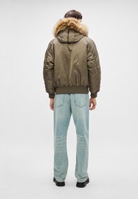 Osoba ubrana w oliwkową bombkę z kapturem wykończonym futrem, jasnoniebieskie dżinsy i czarne buty, stojąca tyłem do aparatu.