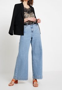 Blazer negro sobre una blusa de patrón de cebra con detalles de encaje, combinado con jeans de pierna ancha azul y sandalias de tacón block naranjas.
