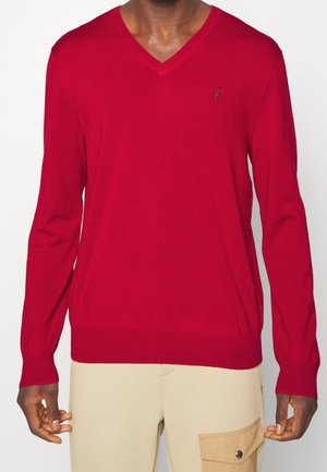 Mężczyzna ubrany w czerwony sweter z dekoltem w serek z długimi rękawami oraz beżowe spodnie z boczną kieszenią, stojący na jednolitym tle.