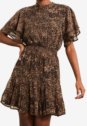 Vrouw die een korte, loszittende jurk met luipaardprint draagt, met gerimpelde mouwen en een geplooide taille, staand met één hand op de heup.