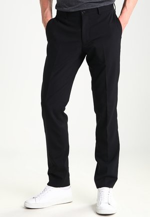 Man draagt strakke zwarte broek en witte sneakers, staand met handen in de zakken tegen een effen witte achtergrond.