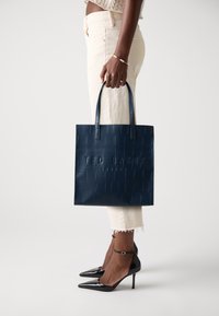 Borsa a spalla blu navy con texture croccante in rilievo, forma piatta e doppi manici, abbinata a scarpe col tacco nere e pantaloni crema.