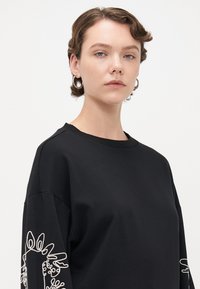 Kvinne med kort brunt hår som har på seg sølv hoop-øreringer og en svart hettegenser med hvit blomsterbrodering på ermene, mot hvit bakgrunn.