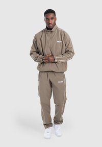 Tuta marrone con accenti bianchi, dotata di giacca con zip e polsini elasticizzati. Abbinata a sneakers bianche e un semplice logo sul petto.