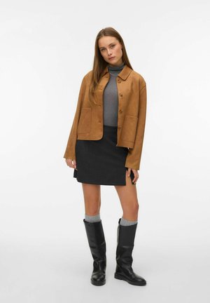 Kvinde iført en brun knapjakke over en grå rullekrave, sort nederdel, grå sokker og sorte knæhøje støvler, stående mod en hvid baggrund.