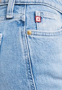 Gaiši zilu džinsa bikšu kabatas ar redzamu šuvumu, mazu sarkanu etiķeti ar baltu kvadrātu un sarkanu zvaigzni, kā arī misiņa kniedes pogu.