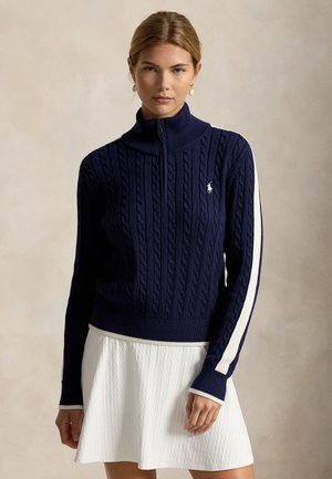 Femme portant un pull en maille torsadée bleu marine avec une bande blanche sur la manche et une jupe blanche texturée sur un fond neutre.