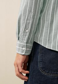 Grøn skjorte med knapper og hvide lodrette striber, med knapmanchetter og en glat tekstur, båret over mørk denim.