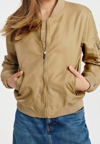 Chaqueta bomber de color marrón claro con cierre de cremallera en la parte delantera, puños acanalados y dos bolsillos laterales, combinada con una falda de mezclilla. Textura suave y brillante.