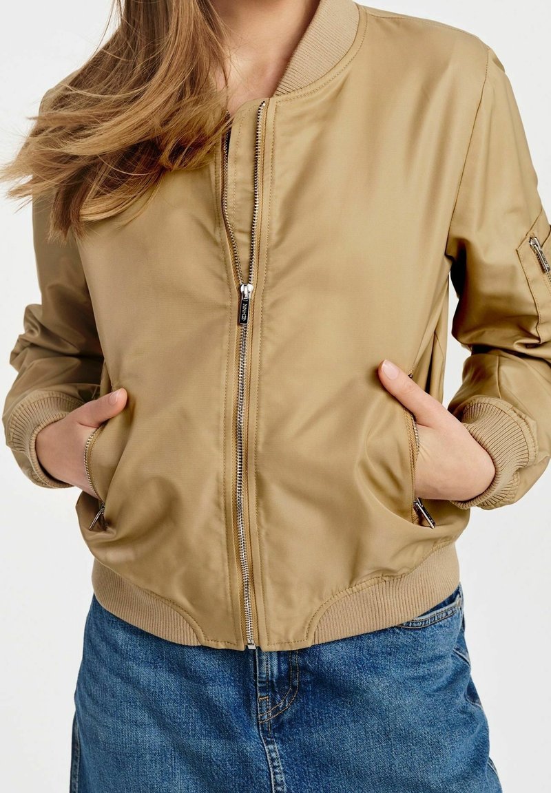 Chaqueta bomber de color marrón claro con cierre de cremallera en la parte delantera, puños acanalados y dos bolsillos laterales, combinada con una falda de mezclilla. Textura suave y brillante.