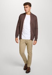 Brun teksturert cardigan med glidelås foran, brukt over en hvit t-skjorte og kombinert med khakifargede bukser. Brune ankelstøvler fullfører antrekket.