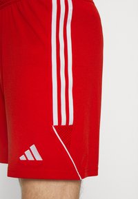 Rode sportieve shorts gemaakt van ademend materiaal, met drie witte verticale strepen aan de zijkant en een klein wit logo aan de zoom.