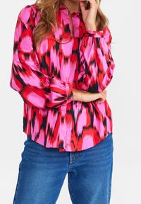 Camicia a maniche lunghe in un vivido rosso, rosa e con motivi astratti neri, con colletto a bottoni e dettagli sui polsini. Il tessuto sembra liscio e leggero.