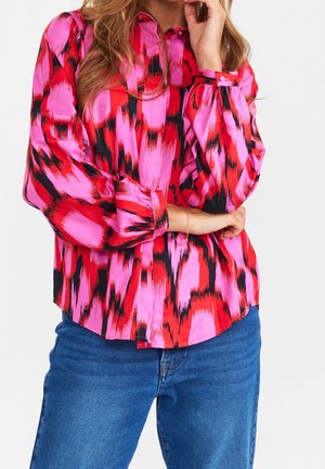 Chemise à manches longues dans des motifs abstraits éclatants de rouge, rose et noir, avec un col à boutons et des détails aux poignets. Le tissu semble lisse et léger.