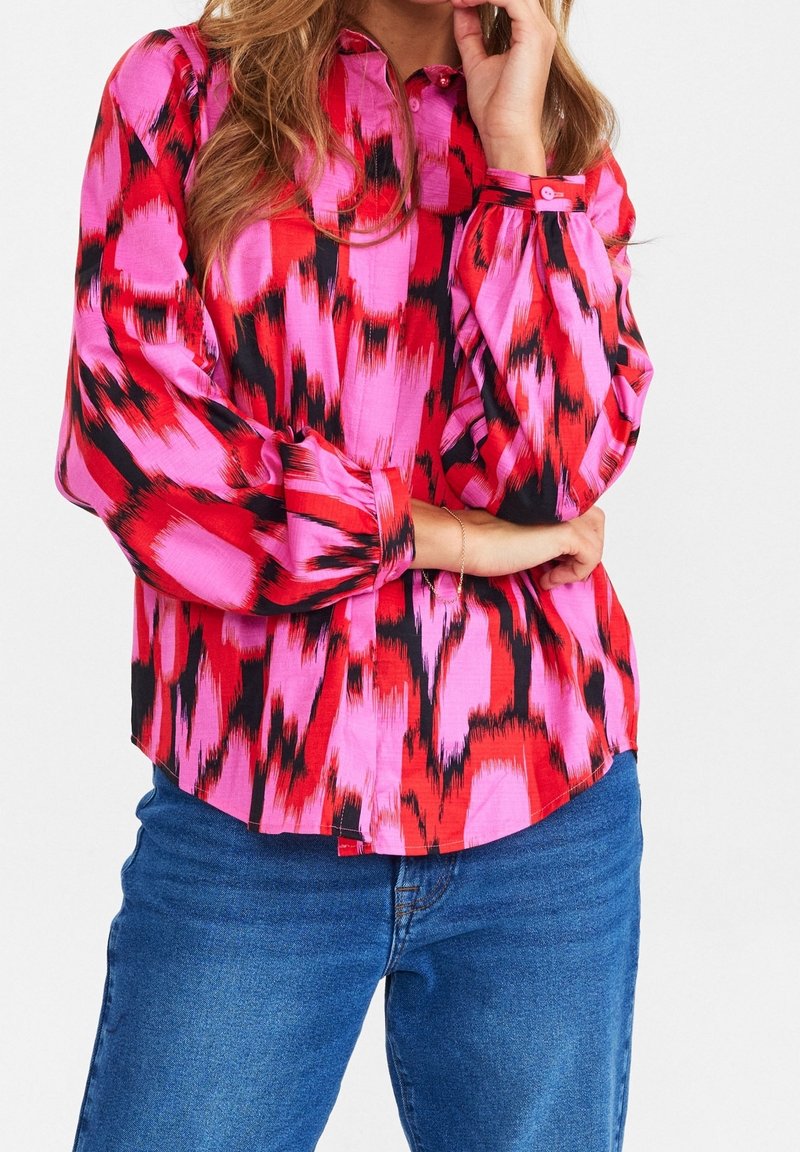 Camicia a maniche lunghe in un vivido rosso, rosa e con motivi astratti neri, con colletto a bottoni e dettagli sui polsini. Il tessuto sembra liscio e leggero.