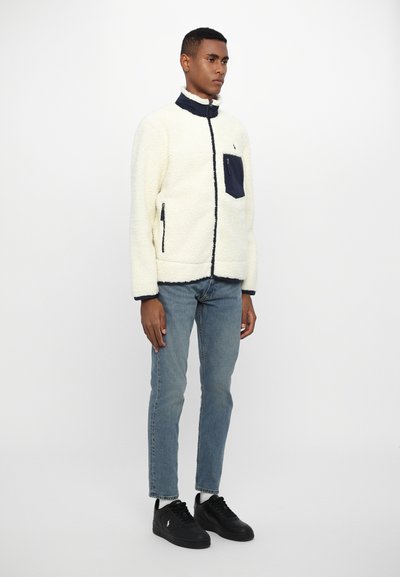 Fuzzy cream jacket with navy accents, high collar, and zip pockets, paired with light blue jeans and black sneakers. Smooth texture.