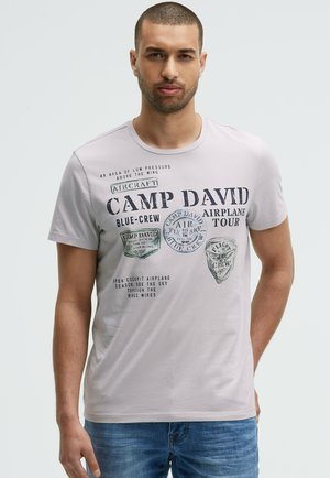 Mann mit kurzen Haaren, der ein hellgraues Camp David T-Shirt mit luftfahrtthematischem Text und blaue Jeans trägt, steht vor einem schlichten Hintergrund.