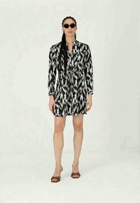 Frau in einem schwarz-weiß gemusterten Kleid, mit dunkler Sonnenbrille und braunen Absatzsandalen, stehend vor einem schlichten Hintergrund.