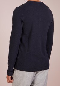 Navy long-sleeve shirt in textured fabric with a round neckline and side slits, paired with light grey pants, viewed from the back.