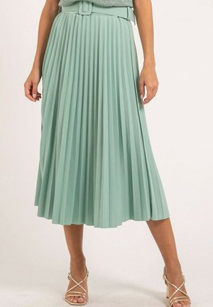 Femme portant une jupe midi plissée taille haute vert clair avec une ceinture assortie et des sandales à talons hauts dorées à lanières.