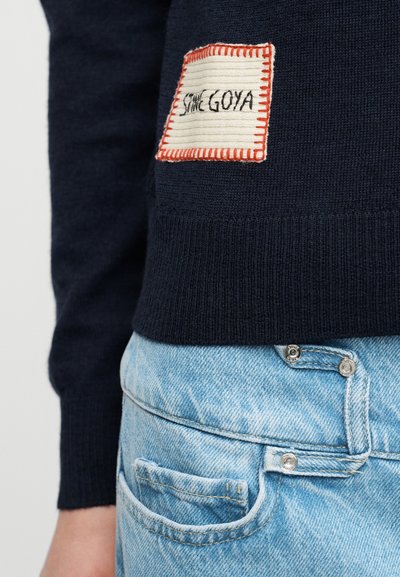 Μπλε πλεκτό πουλόβερ με ορθογώνιο μπεζ μπαλώμα πλαίσιο σε πορτοκαλί, με την επιγραφή "STONE GOYA", φορεμένο πάνω από ανοιχτό μπλε τζιν.