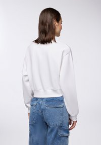 Felpa bianca con design corto, polsini a coste e colletto rotondo, abbinata a jeans cargo blu con molteplici tasche e una texture sbiadita.