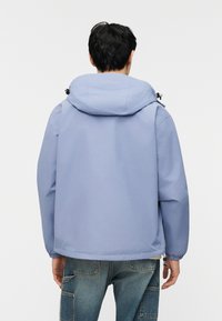 Άτομο που φοράει ανοιχτό μπλε μπουφάν με κουκούλα και ελαστικά μανίκια, όρθιο με την πλάτη προς την κάμερα, συνδυασμένο με μπλε τζιν παντελόνι.