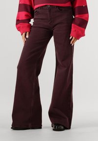 Vrouw in wijde, donkere paarse broek, gecombineerd met een rode en zwarte gestreepte lange mouwen top en zwarte schoenen, staande op een effen achtergrond.