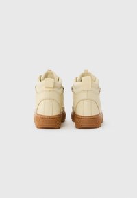 Et par beige mid-top sneakers med tykke brune såler sett fra bak på en hvit bakgrunn.