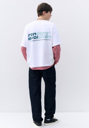 Jeune homme portant un t-shirt blanc par-dessus une chemise à manches longues rouge rayée, un jean foncé et des baskets noires, face à un mur blanc.