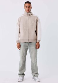 Joven vestido con un suéter beige y blanco con cremallera, jeans gris claro y zapatillas blancas, de pie contra un fondo limpio y claro.