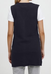 Der marineblaue Strick-Westen verfügt über ein strukturiertes Muster, ärmelloses Design und einen abgerundeten Saum, kombiniert mit einem weißen T-Shirt und grauen Hosen.