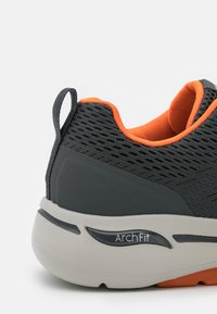 Scarpa sportiva in mesh nera con accenti arancioni e suola grigia che presenta il marchio "ArchFit". Include un'etichetta per indossarla facilmente.