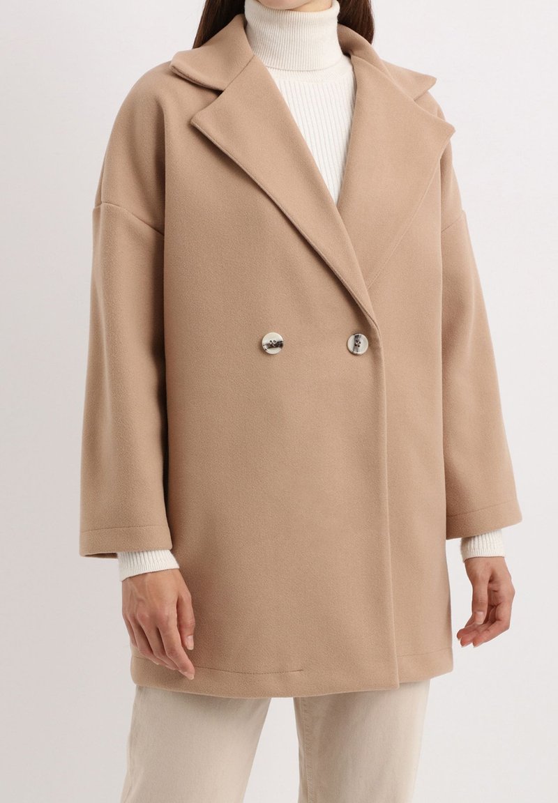 Abrigo de mezcla de lana beige, de doble botonadura, con amplios solapas, botones en tono plateado y corte relajado. Mangas anchas cubren un cuello de tortuga blanco de canalé.