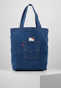Sac fourre-tout en denim avec deux poignées robustes, arborant un petit motif Hello Kitty qui émerge d'une poche arrière. Tissu bleu avec une finition texturée.