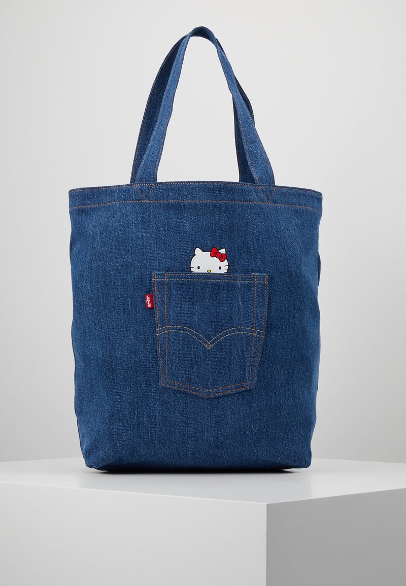 Sac fourre-tout en denim avec deux poignées robustes, arborant un petit motif Hello Kitty qui émerge d'une poche arrière. Tissu bleu avec une finition texturée.