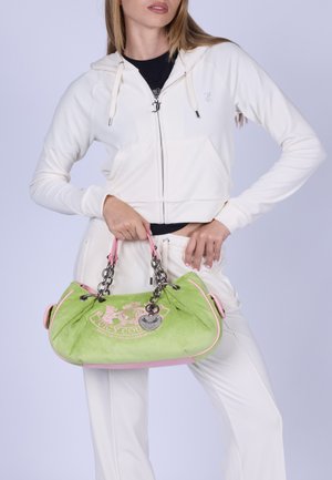 Donna con tuta bianca che tiene una borsa Juicy Couture verde e rosa con tracolle a catena e dettaglio logo.