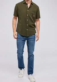 Camicia verde oliva con maniche corte e bottoni, dotata di taschino frontale, abbinata a jeans blu e sneaker bianche, per un look casual.