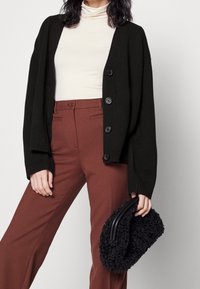 Mujer con cárdigan negro abotonado, camiseta de cuello alto blanca, pantalones color óxido, sosteniendo un bolso negro peludo con asas redondas.