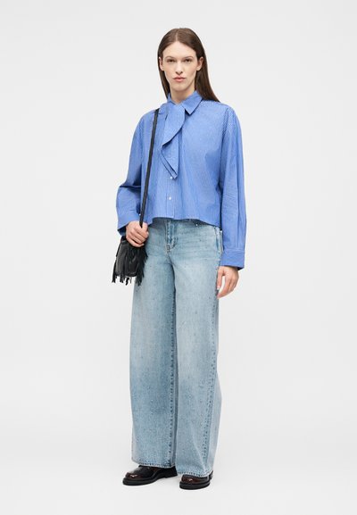 Femme portant un chemisier à rayures bleues avec un détail de cravate au col, un jean large bleu clair orné, des chaussures noires, et portant un sac noir à franges.