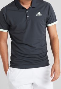 Homme portant un polo Adidas noir avec des bordures blanches aux manches et un short blanc, les mains détendues le long des hanches sur un fond uni.