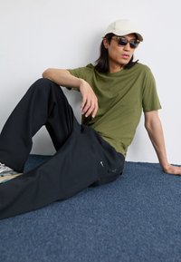 Grünes Baumwoll-T-Shirt, schwarze lässige Hose mit Reißverschlussdetails, beiger Hut und Sonnenbrille, sitzend auf einem blauen Teppich vor einer weißen Wand.