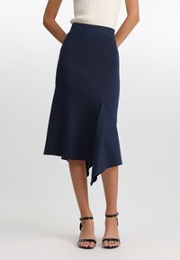 Marineblå asymmetrisk skjørt i stretchy stoff med flagrende kant, med en sideåpning og en jevn midjebånd. Kombinert med svarte sandaler med rem.