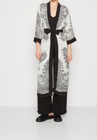 Long white robe with black paisley print, black cuffs, and tie belt. Paired with black wide-leg pants and black shoes.
