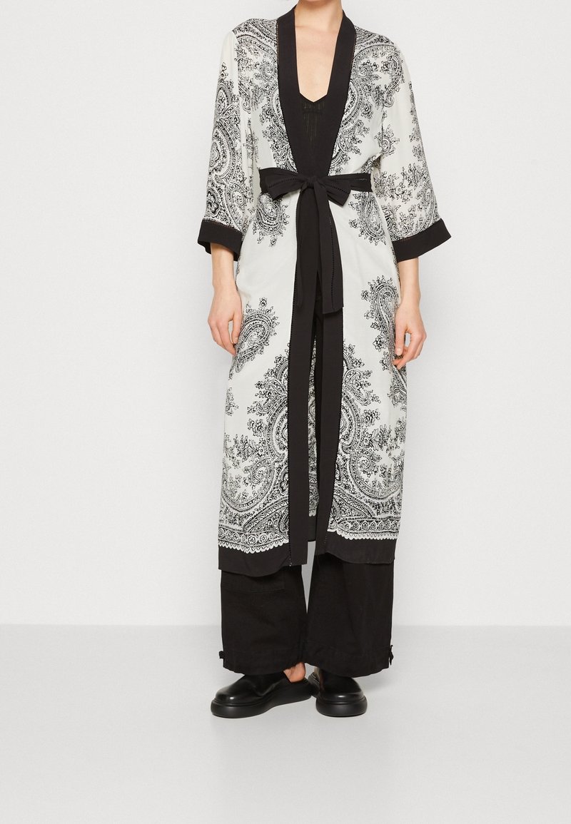 Long white robe with black paisley print, black cuffs, and tie belt. Paired with black wide-leg pants and black shoes.