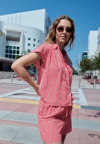 Piros-fehér csíkos ing és rövidnadrág szett, rövid ujjú, galléros, gombos elülső résszel, laza fazon, kör alakú napszemüveggel viselve, kék ég háttérrel.