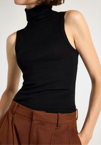 Sort ærmeløs turtleneck top lavet af et glat materiale, parret med højvristede, plisserede brune bukser.