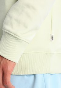 Sweat-shirt vert clair avec poignets et ourlet côtelés, comportant une petite étiquette logo sur le côté. Texture douce, coupe décontractée, et design minimaliste.