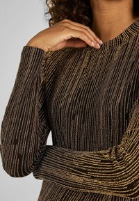 T-shirt à manches longues avec un motif rayé noir et or, doté d'un tissu texturé et d'un col rond, avec un détail froncé sur les manches.