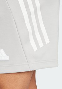 Ljust grå Adidas-shorts med tre vita ränder och logotyp på låret, bärs på en persons ben.